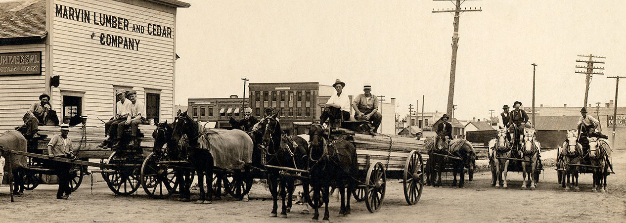 Historical photo of Marvin Lumber company employees: men gather on horse-drawn wagons carrying lumber outside of the Marvin Lumber Company building