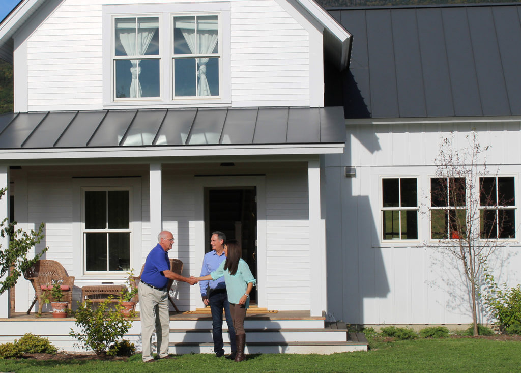 A salesman shakes the hands of a husband and wife as they stand outside their modern-looking home that has just had new windows and doors installed