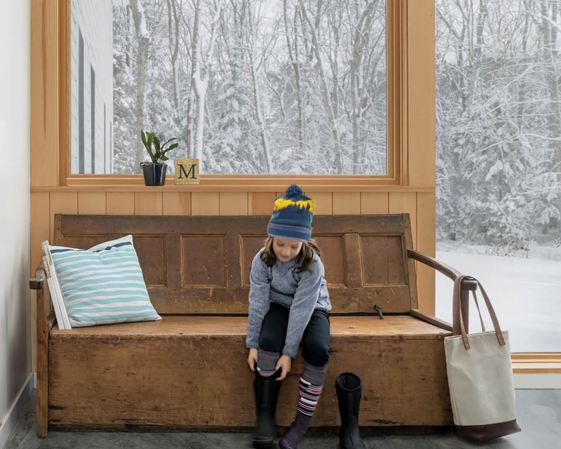 Little girl in mudroom with large glass windows putting on boots to go play in the snow