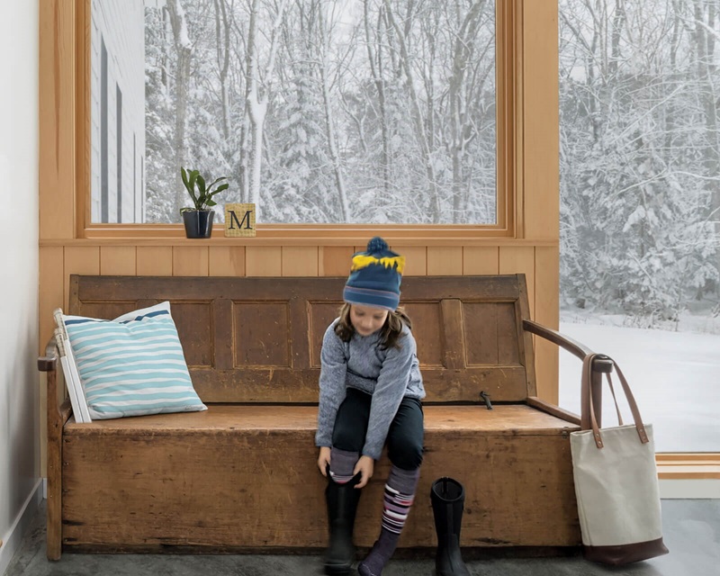 Little girl in mudroom with large glass windows putting on boots to go play in the snow