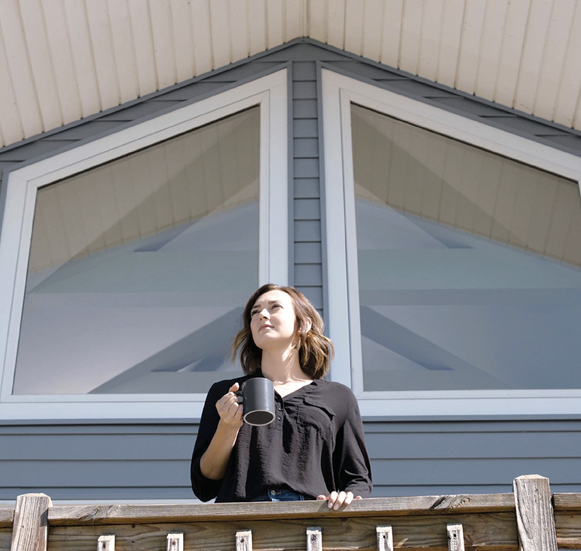 Woman holding a coffee mug and looking out from balcony of gray house with windows